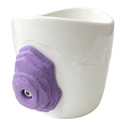 Side view: An off-white ceramic climbing mug, featuring a handle shaped like a 3D purple rock-climbing hold.