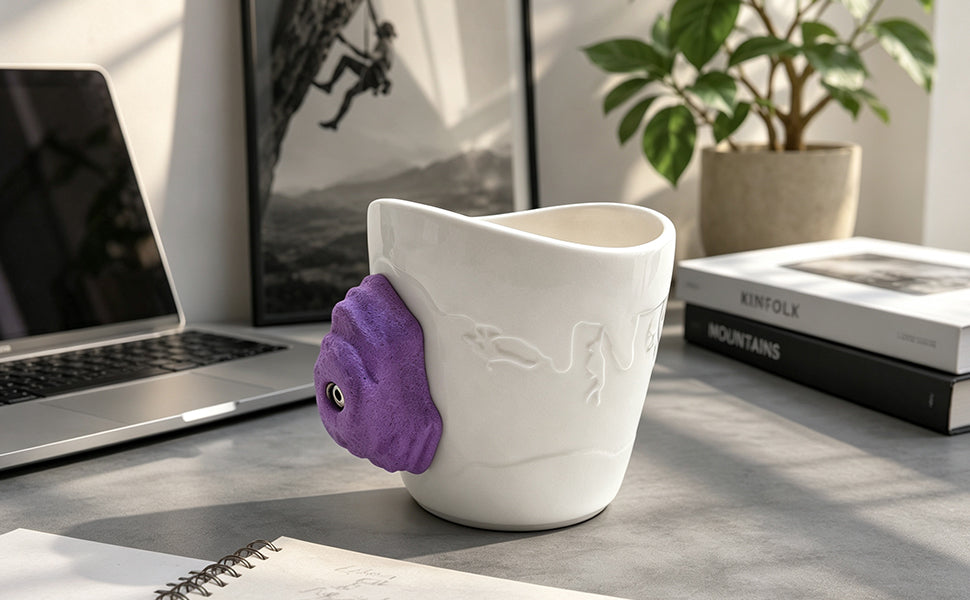 White ceramic rock climbing mug with 3D climbing hold handle on a wooden table by a window