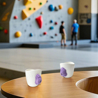 A rock-climbing mug, featuring a handle designed to look like a 3D climbing hold, sits on a table at a bouldering gym.